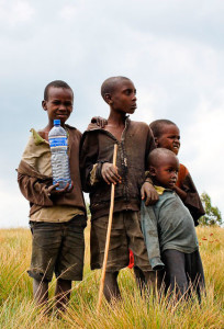 Four children stand together wearing worn clothes. One child is holding a large bottle of clean water.